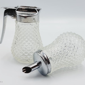 Op de afbeelding: Twee heldere glazen siroopdispensers met een diamantpatroon. Een dispenser heeft een zilveren tuit en een handvat. De andere dispenser heeft een zilveren deksel en een zilveren tuit.