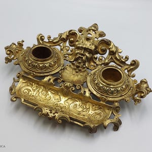 Puede incluir: Un tintero de metal dorado con detalles ornamentados y dos tinteros. El tintero tiene un elemento decorativo central con una cara y un diseño de rollo.