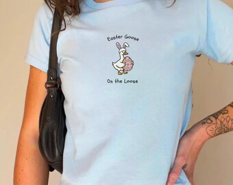 Może przedstawiać: Jasnoniebieski t-shirt z grafiką przedstawiającą białą gęś w różowych króliczych uszach, trzymającą kolorowe jajko wielkanocne we wzory. Tekst nad gęsią brzmi "Easter Goose", a tekst pod gęsią brzmi "On the Loose."