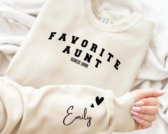 Może przedstawiać: Beżowa bluza z okrągłym dekoltem, posiadająca na piersi czarny napis "FAVORITE AUNT SINCE 1999" czcionką typu college oraz imię "Emily" wraz z dwoma małymi czarnymi sercami nadrukowanymi na rękawie pismem odręcznym.