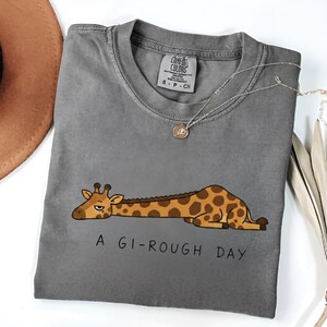 Może przedstawiać: Szara koszulka Comfort Colors z kresk&oacute;wkową żyrafą w brązowe plamy, leżącą. Pod żyrafą wydrukowano napis "A GI-ROUGH DAY". Srebrny naszyjnik z małą zawieszką leży na koszulce.