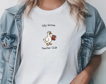 Może przedstawiać: Biała koszulka z kreskówkową gęsią trzymającą książkę, z napisem "Silly Goose Teacher Club". Gęś jest biała z pomarańczowymi stopami i żółtym dziobem. Koszulka jest noszona z dżinsową kurtką.
