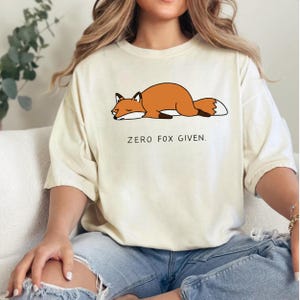 May include: Cream-colored t-shirt featuring a cartoon fox in shades of orange, brown, and white, lying down. The text "ZERO FOX GIVEN" is printed below the fox in black, sans-serif font. The shirt is made of a soft material.