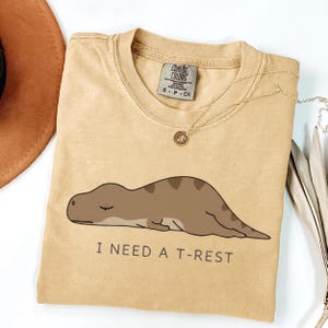 May include: Mustard-colored t-shirt featuring a cartoon dinosaur lying down. The text "I NEED A T-REST" is printed below the dinosaur. The shirt has a crew neck and short sleeves. A gold necklace is draped on the shirt.