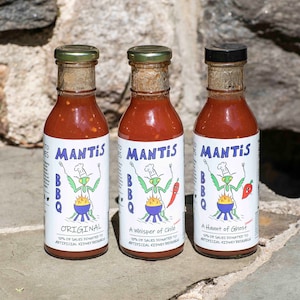 Puede incluir: Tres botellas de vidrio de salsa BBQ Mantis con diferentes perfiles de sabor: Original, Un susurro de chile y Un toque de fantasma. Cada botella presenta un chef mantis de dibujos animados con una parrilla azul y amarilla.
