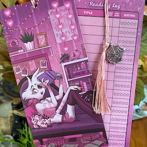 May include: Two purple bookmarks with a pink tassel and a silver spiderweb charm. One bookmark features a cartoon illustration of a character reading, the other has a reading log. The illustration includes the text "Picture Perfect".