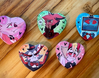 May include: Five heart-shaped badges with cartoon character designs. The badges feature various characters in pink, green, and blue, with detailed illustrations and bold outlines. The badges are arranged on a wooden surface.