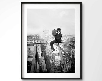 May include: A black and white photograph of a man sitting on a metal beam high above a city. He is wearing a suit and a hat and is holding a camera. The city is in the background and the buildings are tall and narrow.