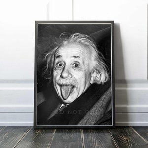May include: Black and white framed portrait of Albert Einstein sticking out his tongue. The image captures Einstein's expressive face, with his eyes wide and his tongue extended. The portrait is set against a white wall and dark wood floor.