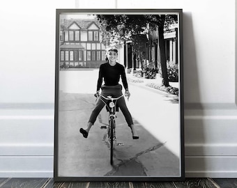 Op de afbeelding: Een zwart-witfoto van een vrouw die op een fiets rijdt op een straat. Ze draagt een zwarte turtleneck trui en broek. De straat is omzoomd met huizen en bomen.