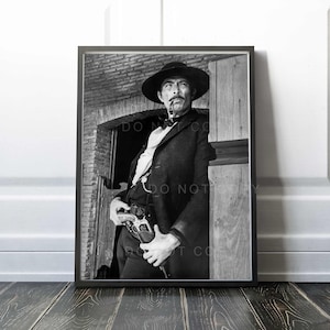 Op de afbeelding: Een zwart-witfoto van een man in een cowboyhoed en pak, die een pistool vasthoudt en een pijp rookt. De man staat in een deuropening en kijkt naar de zijkant. De tekst "Do Not Copy" is zichtbaar op de afbeelding.