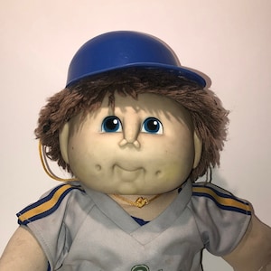 Puede incluir: Una muñeca Cabbage Patch Kids con una gorra de béisbol azul, una camiseta de béisbol gris y amarilla con el texto "Cabbage Patch Kids" en la parte delantera y zapatillas blancas con detalles amarillos.