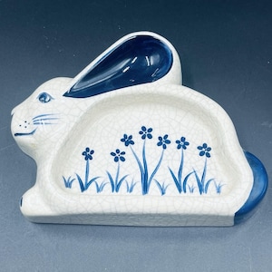 May include: White ceramic bunny-shaped dish with blue accents. The dish features a cracked glaze finish, a blue-painted ear, and a blue tail. Blue floral design decorates the interior of the dish, adding a touch of spring.