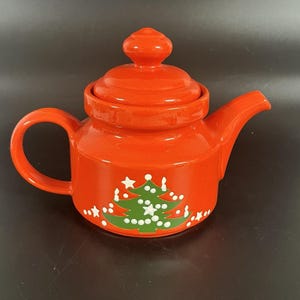 May include: A festive, red ceramic teapot with a matching lid and handle. The teapot features a green Christmas tree design with white star and dot accents. A charming holiday-themed kitchenware item.