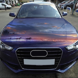 Op de afbeelding: Een donkerblauwe auto met een motorkap met een afbeelding van paarden die bij zonsondergang over water rennen. De grille en koplampen van de auto zijn zichtbaar.