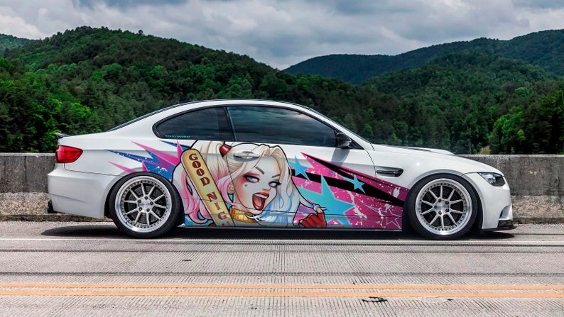 May include: White sports car with custom graphics featuring a cartoon character with the text "GOOD NIGHT" on a baseball bat. The car has a black spoiler and silver rims, parked on a road with a mountain backdrop.