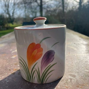 May include: A ceramic jar with a lid, featuring a floral design. The jar is painted with orange and purple flowers with green stems and leaves. The lid has a small, round handle with an orange top.