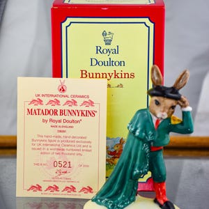 Puede incluir: Una figura de Royal Doulton Bunnykins de un conejo vestido de matador, con una capa verde y pantalones rojos. La figura está sobre una base de color crema, con una caja roja y amarilla y una tarjeta con texto en el fondo.
