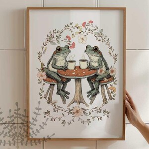 Peut inclure: Deux grenouilles vertes assises à une petite table en forme de champignon, buvant dans des tasses. Elles sont entourées d'une couronne de fleurs et de feuilles.