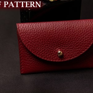 Può includere: Un piccolo portafoglio in pelle color borgogna con chiusura a lembo e un bottone dorato. Il portafoglio ha una superficie testurizzata e granulosa.