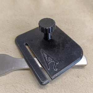 Può includere: Una piastra per fotocamera nera stampata in 3D con una piccola manopola in cima. La piastra ha una fessura per una barra di metallo e il testo "AC" è visibile sulla piastra.