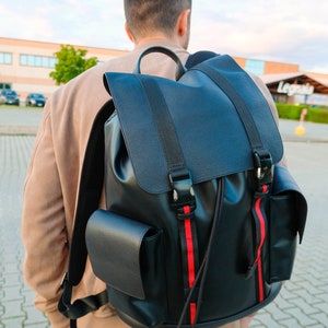 Può includere: Uno zaino in pelle nera con strisce rosse e bianche. Lo zaino ha due grandi tasche sul davanti e una chiusura con coulisse.
