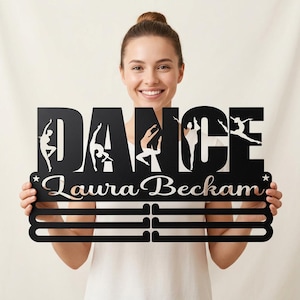 May include: Black metal dance medal holder with the word "DANCE" cut out, featuring dancer silhouettes. The name "Laura Beckham" is also cut out. Includes three horizontal bars for displaying medals. A star is on each side.