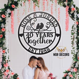 Puede incluir: Un letrero de metal negro con el texto "Rose & Jorden 40 Years Together Est 1985" y un dise&ntilde;o de dos p&aacute;jaros en una rama con corazones y flores. El letrero est&aacute; colgado en una pared con una pareja de pie frente a &eacute;l.