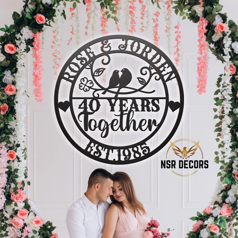 Puede incluir: Un letrero de metal negro con el texto "Rose & Jorden 40 Years Together Est 1985" y un dise&ntilde;o de dos p&aacute;jaros en una rama con corazones y flores. El letrero est&aacute; colgado en una pared con una pareja de pie frente a &eacute;l.