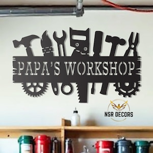Puede incluir: Arte de pared de metal negro con un letrero que dice "Papa's Workshop". El letrero está decorado con varias herramientas, incluyendo un martillo, una sierra, una llave y unos alicates. El arte está montado en una pared blanca.