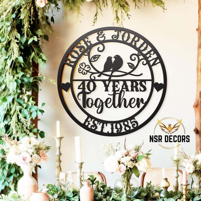 Puede incluir: Un letrero redondo de metal negro con los nombres "Rose & Jordan" y el texto "40 Years Together Est. 1985". Dos p&aacute;jaros est&aacute;n posados en una rama con hojas y flores. El letrero est&aacute; colgado en una pared con decoraciones florales.