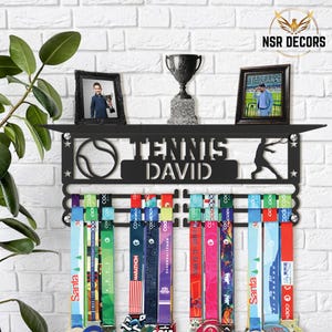 Puede incluir: Expositor de medallas de metal negro con una pelota de tenis y una silueta de un jugador de tenis. El expositor tiene un estante con el nombre "TENNIS DAVID" y espacio para medallas. El expositor está montado en una pared de ladrillo blanco.