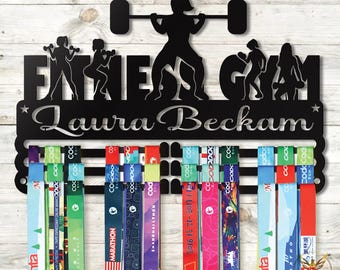 May include: Black metal fitness-themed medal display with the words "Fitness Gym" and the name "Laura Beckham". The display features silhouettes of weightlifters and runners, with multiple ribbons hanging below.
