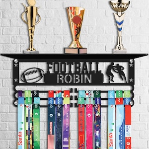 May include: A black metal medal display rack with the words "Football Robin" on it. The rack has six rows of hooks for hanging medals. There are several colourful medals hanging on the hooks. There are three gold trophy cups on the top shelf.