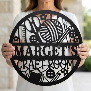May include: A circular black metal wall sign featuring a cutout design of craft supplies including scissors, a yarn ball, buttons, a ruler, and pencils. The center horizontal bar displays the text MARGET'S CRAFT ROOM in a bold, sans-serif font.