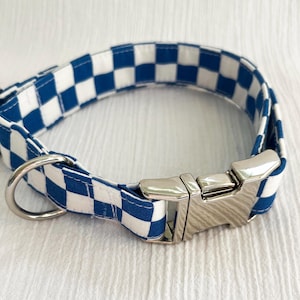 May include: A blue and white checkered dog collar with a silver metal buckle and D-ring. The collar is made of fabric and is designed to be worn around a dog's neck. The pattern is a classic checkerboard design.