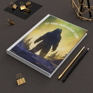 May include: A white journal with a colorful illustration of a Bigfoot standing in a snowy forest. The text "Let your imagination flow" is printed on the cover.
