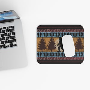 May include: A rectangular mousepad with a Bigfoot theme. The design features a Bigfoot silhouette, pine trees, and footprints on a patterned background of brown, blue, and gold. A white computer mouse is on the right side.