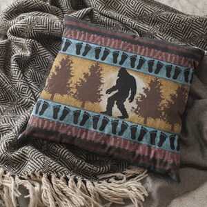 May include: Square decorative pillow featuring a Bigfoot silhouette in a forest scene. The pillow has a tan background with brown trees and a Bigfoot figure. The design includes blue and maroon borders with black footprints.