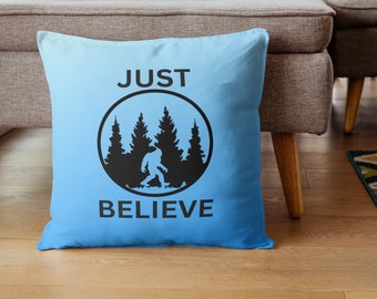 Może przedstawiać: Niebieska poduszka ozdobna z napisem "JUST BELIEVE" w czarnych literach. Czarna sylwetka Bigfoota idącego przez las znajduje się w kółku nad tekstem.