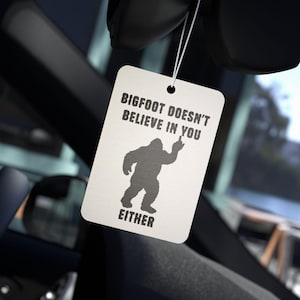 May include: A rectangular car air freshener with the text "BIGFOOT DOESN'T BELIEVE IN YOU EITHER" and a silhouette of Bigfoot giving the middle finger. The air freshener hangs from a white string.