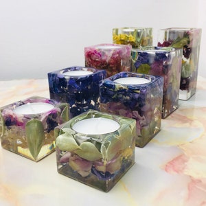 May include: A selection of square resin candle holders, each containing dried flowers in purple, blue, and green hues. Each holder features a white tea light candle. The candle holders are displayed on a light-coloured surface.