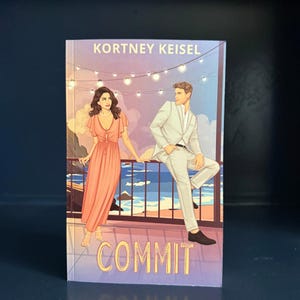 May include: A paperback book titled COMMIT by Kortney Keisel. The cover features an illustration of a person in a peach-colored dress and a person in a light-colored suit standing on a balcony overlooking the ocean under string lights.
