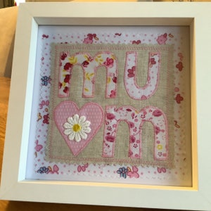 May include: A framed piece of fabric art with the word "mum" spelled out in pink and white fabric letters with a pink heart and a white daisy in the centre. The background is a pink and white polka dot fabric.