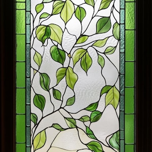 Könnte beinhalten: Buntglasfenster mit einem Design aus grünen Blättern und Ranken. Die Blätter sind in verschiedenen Grüntönen gehalten, mit schwarzen Umrissen. Das Fenster ist mit einem dunkelbraunen Rahmen und einem grünen Rahmen mit rechteckigen Blöcken eingefasst.