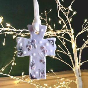May include: A pale lilac cross-shaped decoration adorned with small flowers and white pearl details, suspended by a white ribbon. The ornament hangs in front of a decorative tree with warm white LED lights, creating a soft, illuminated backdrop.