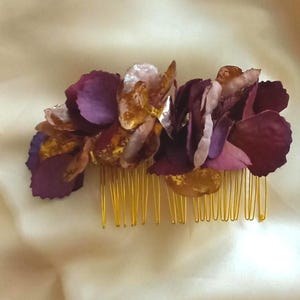 May include: A hair comb featuring a cluster of layered petals in shades of deep purple, light lavender, and metallic gold. The decorative floral arrangement is attached to a gold-toned metal comb with multiple thin, parallel teeth.