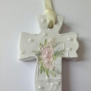 May include: A white, decorative cross with a cream-colored ribbon for hanging. The cross features a floral design with pink and green accents, along with small, pearl-like embellishments. The cross is approximately 4 inches in height.