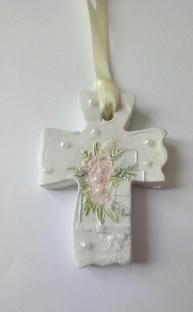 May include: A white, decorative cross with a cream-colored ribbon for hanging. The cross features a floral design with pink and green accents, along with small, pearl-like embellishments. The cross is approximately 4 inches in height.