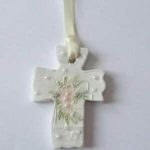 May include: White ceramic cross pendant with a floral design in pink and green. The cross has a ribbon for hanging and is decorated with small, raised dots. A delicate religious ornament.